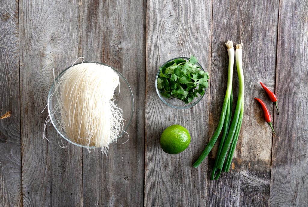 A bowl of dry rice noodles, cilantro, lime and green onions.