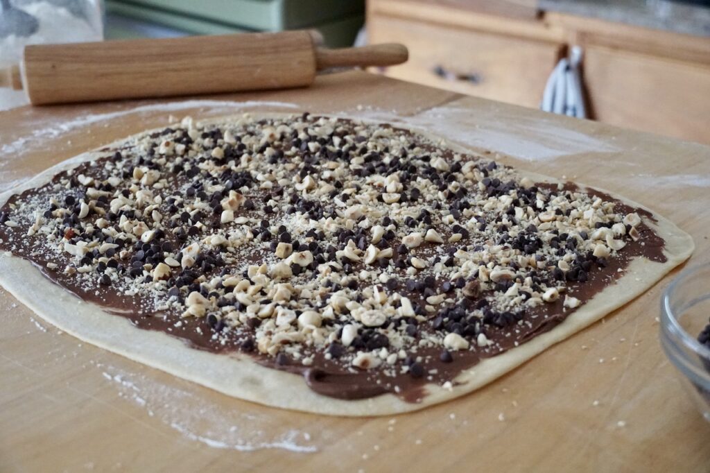 A large rectangle of dough smeared with the spread and sprinkled with chopped hazelnuts and chocolate chips.
