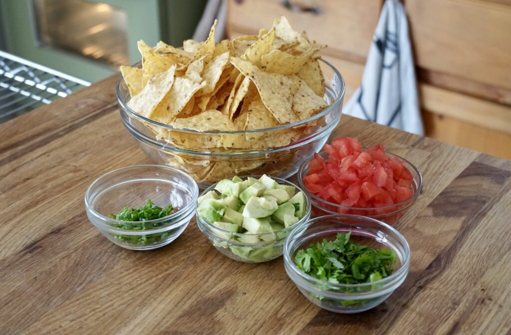 Garnishes for the dip include; avocado, green onions, cilantro and tomatoes served with crispy corn tortilla chips.