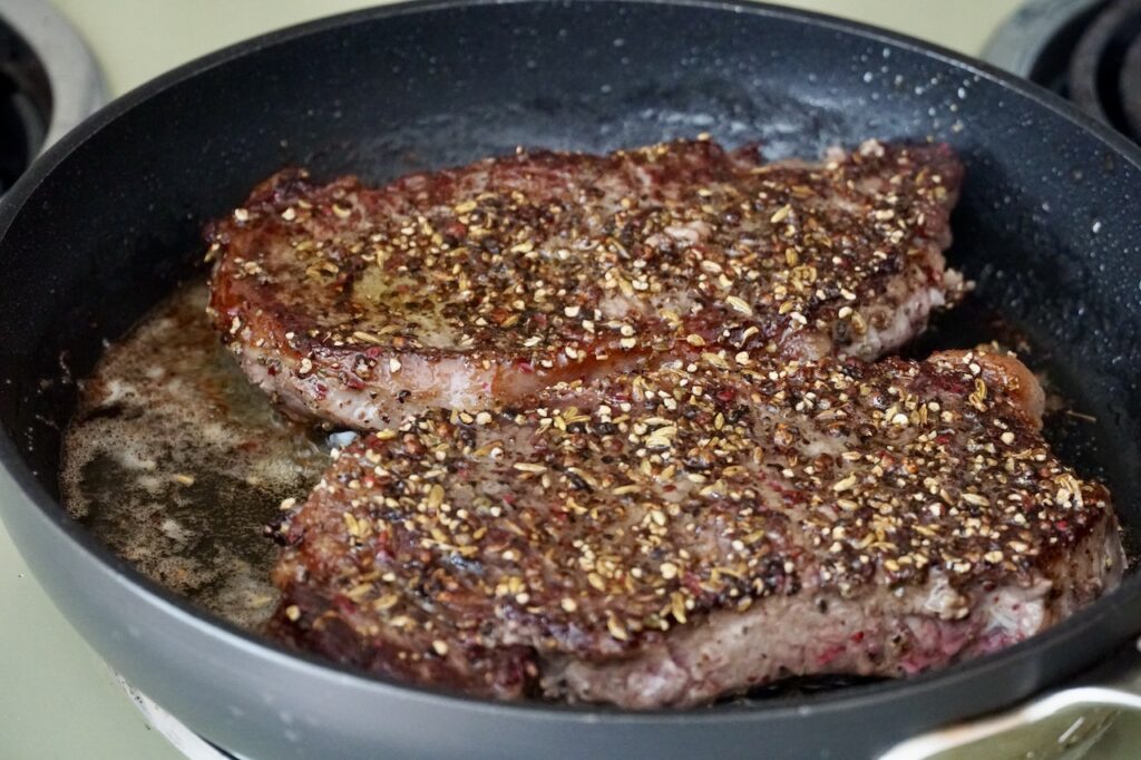 The sirloin strip steaks sizzling in a hot skillet on stovetop.