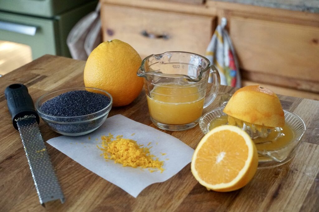 A navel orange cut in half, juiced and zested next to poppy seeds.