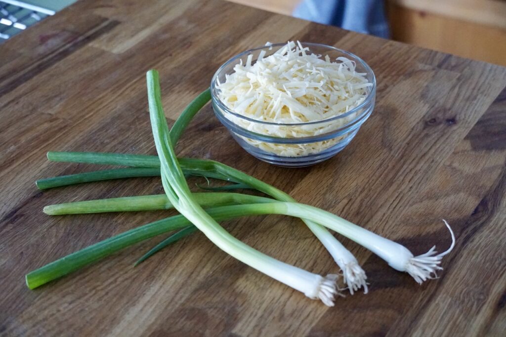 A bowl of grated Gruyere cheese sitting next to three green onions.