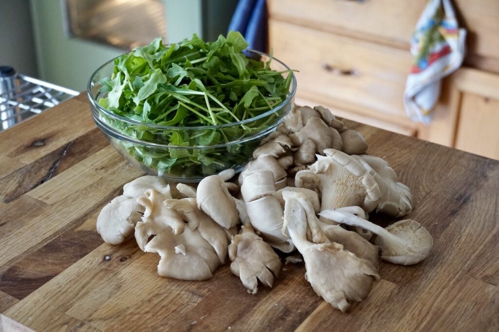 A bowl of baby arugula next to bunches of oyster mushrooms.