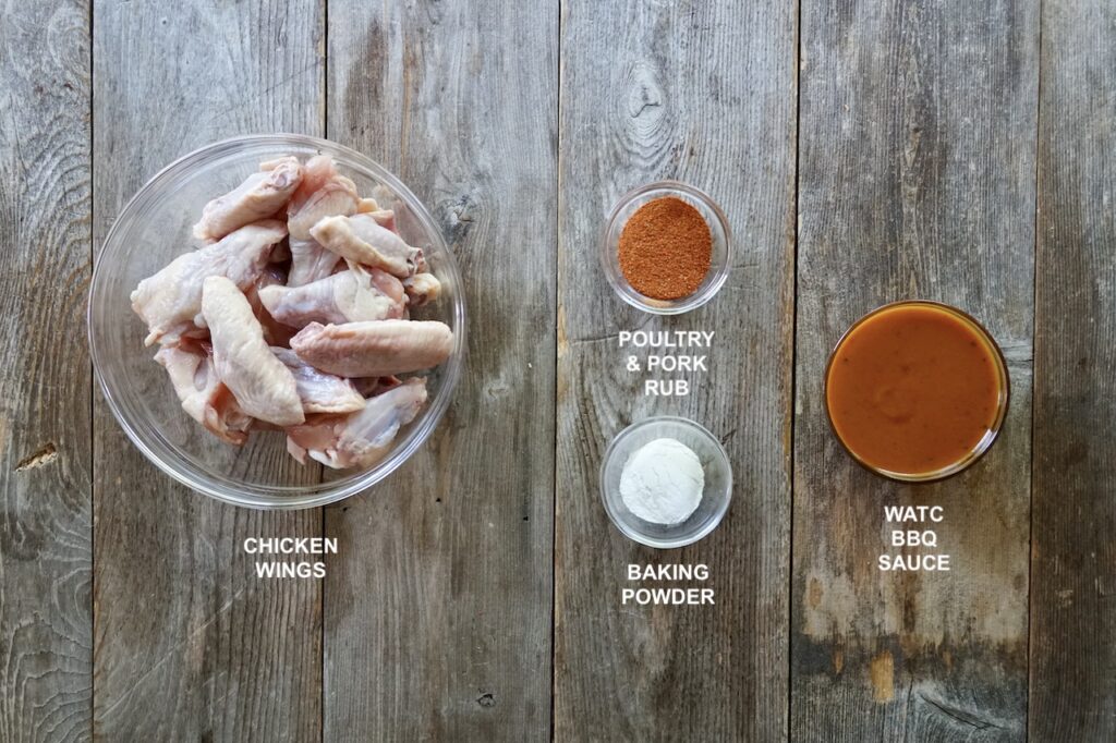 The ingredients for the recipe including a bowl of raw chicken wings, baking powder, poultry and pork rub and tangy barbecue sauce.