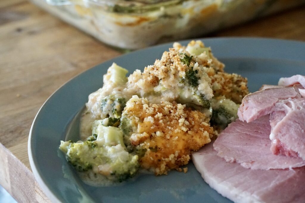 Cheesy Broccoli Casserole served on a dinner plate with oven-baked ham.