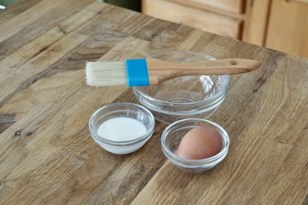 A bowl with an egg and another with whole milk, a pastry brush to the side.