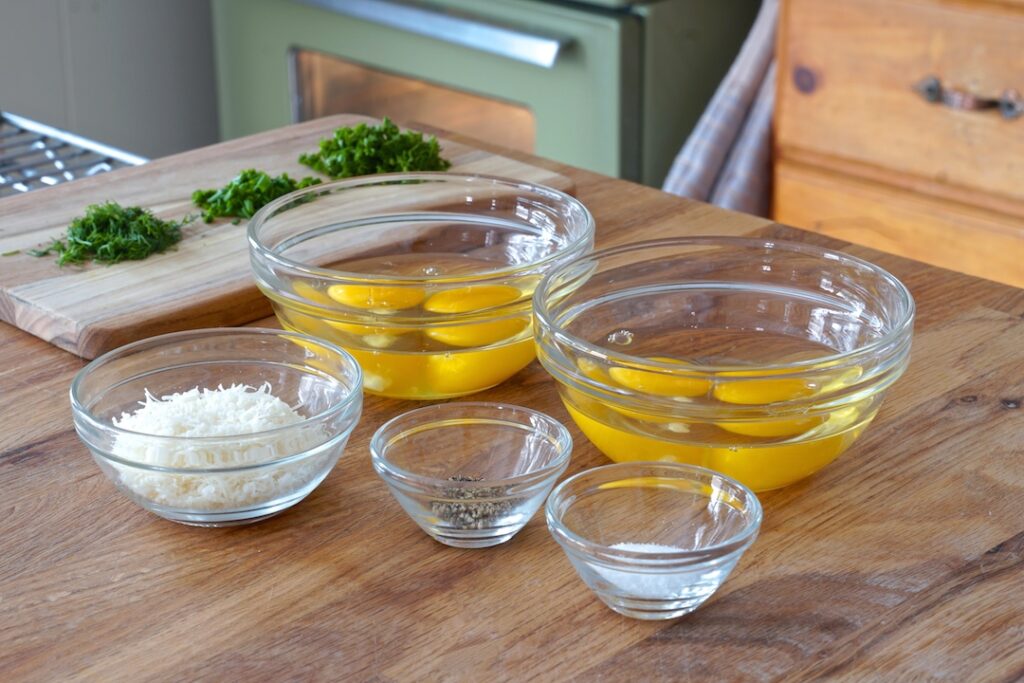 Eggs separated into bowls, sitting next to salt, pepper, Parmesan and chopped fresh herbs.