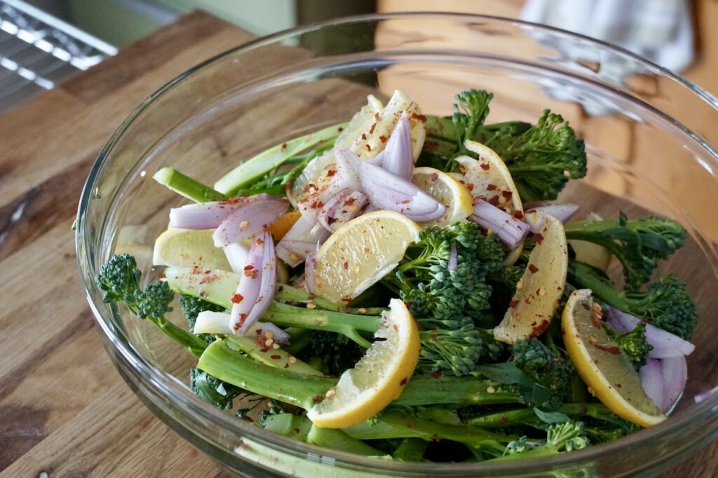 A large glass bowl filled with the broccolini, lemon wedges and sliced shallots.