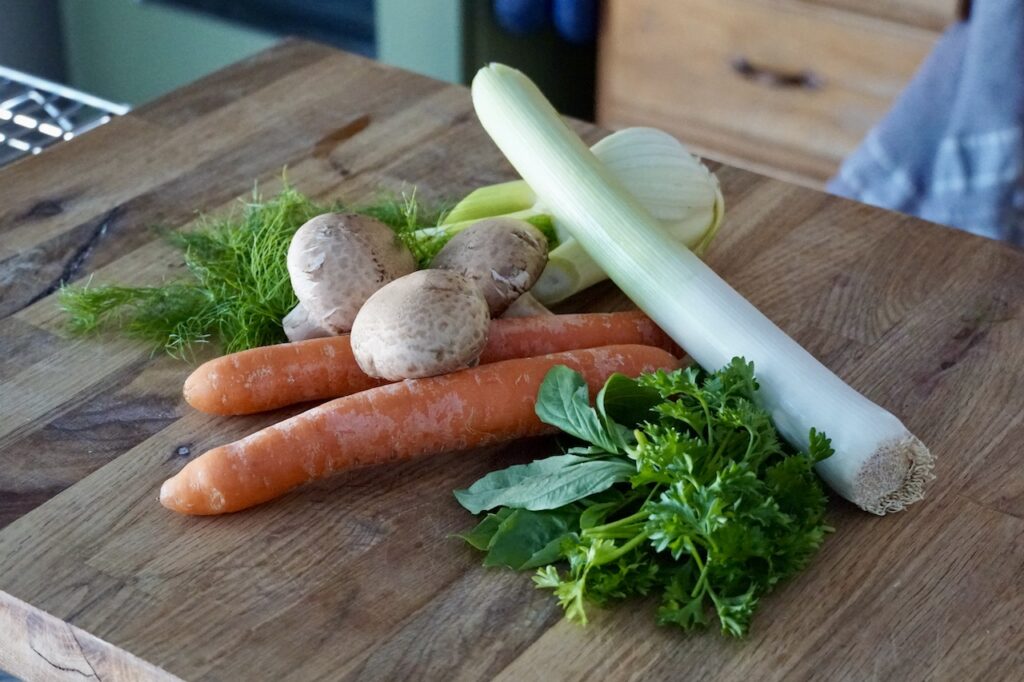 Carrots, leek, cremini mushrooms, fennel, parsley and basil sitting on a wooden cutting board.