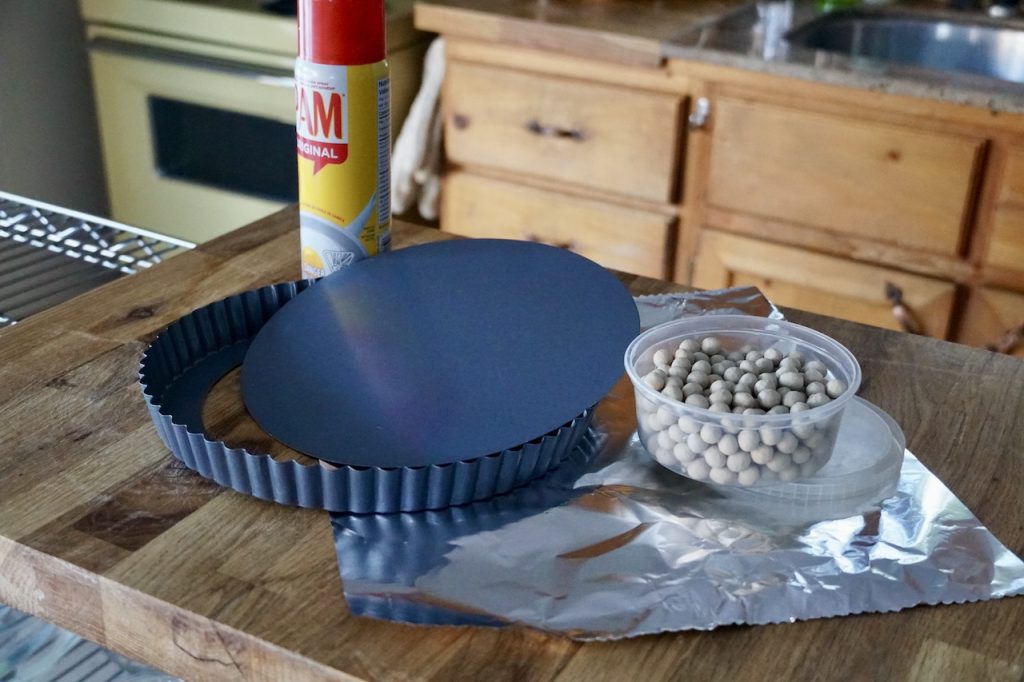 A fluted metal tart pan with a removable bottom, a square of foil and a container of clay pie weight.