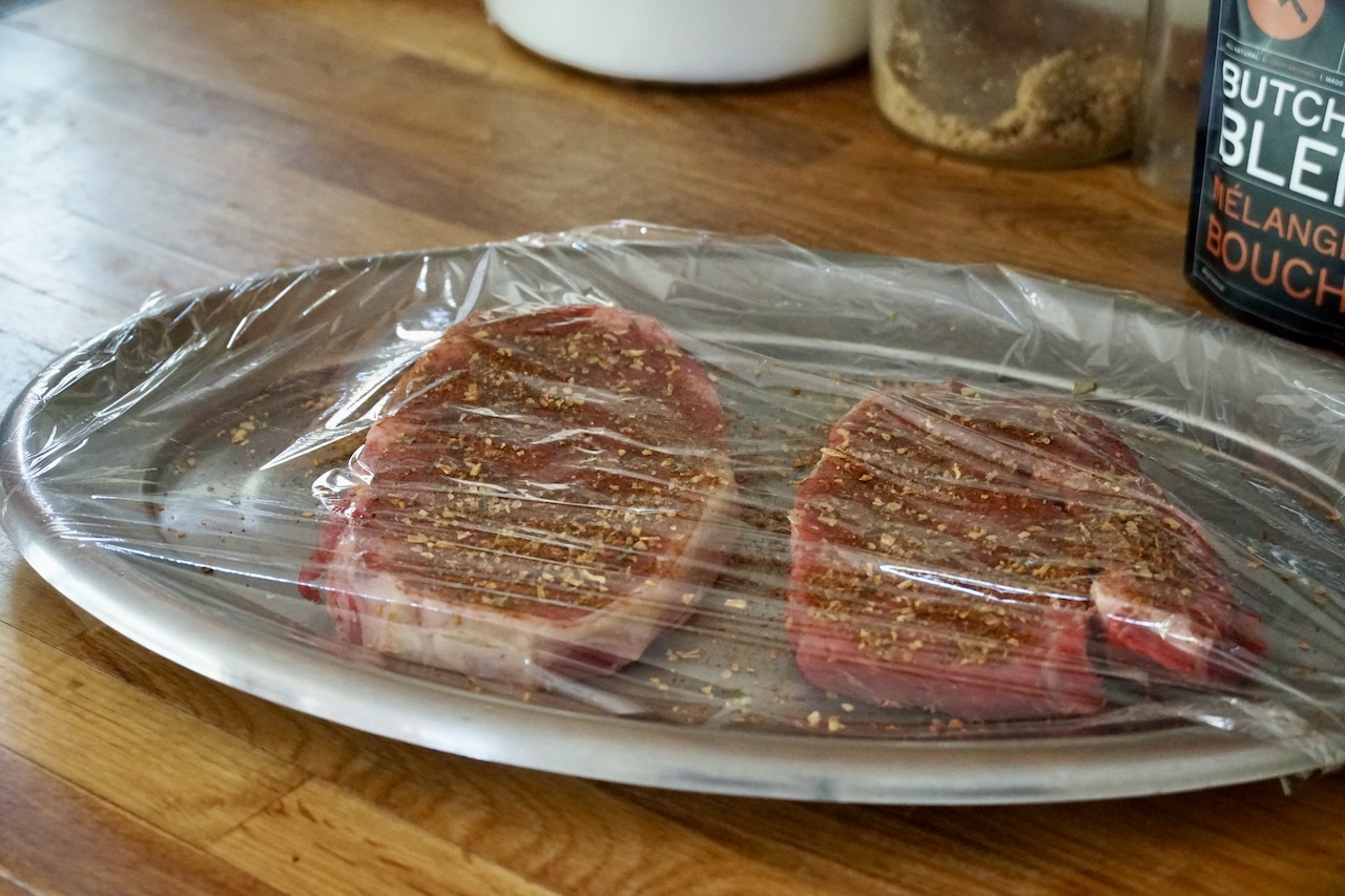 The seasoned ribeye steaks covered, resting on the counter as the steaks come to room temperature.