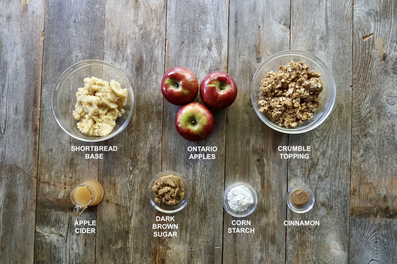 Ingredients for the apple pie crumble bars, assembled in small bowls include: Ontario apples, the topping mixture, the shortbread base plus, dark brown sugar, corn starch, apple cider, and cinnamon.
